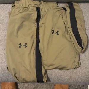 Under Armour Tracksuit Olive Green - Säljer en olivegrön tracksuit från Under Armour med svarta ränder längs benen och ärmarna. Perfekta för gymmet eller löprundan.