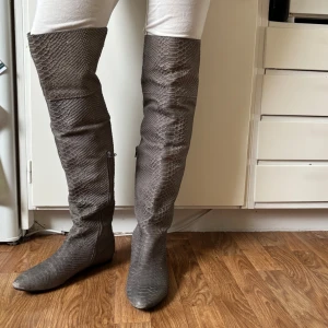 Grå overknee boots med ormskinnsmönster - Säljer ett par grå overknee boots med coolt ormskinnsmönster och rund tå. Skorna har dragkedja på insidan och är tillverkade i syntetmaterial. Aldrig använda, köpt second hand. Så om det syns att de är använda är det från förra ägaren! 