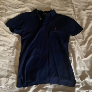 Mörkblå pikétröja från Polo Ralph Lauren - Säljer en mörkblå pikétröja från Polo Ralph Lauren i slim fit-modell. Tröjan har korta ärmar, klassisk krage och röd broderad logga på bröstet. Perfekt för en stilren och sportig look. Andvänd 1-3 gånger max
