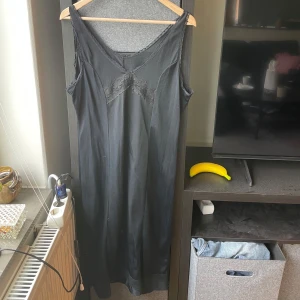 Svart långklänning med spetsdetaljer - Säljer en svart långklänning med v-ringning och fina spetsdetaljer vid halsringningen och bysten. Klänningen har en klassisk och elegant look med ärmlös design och mjukt, glansigt material.