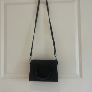 Svart axelväska med handtag - Snygg svart axelväska med justerbar rem och två handtag. Stilren och enkel design som passar till många outfits. Väskan har en fyrkantig form och är perfekt för att bära det viktigaste. PRIS GÅR ATT DISKUTERAS 