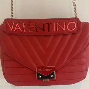 Röd axelväska från Valentino - Säljer en snygg röd axelväska från Valentino med kedjerem och quiltad design. Väskan har ett stort VALENTINO-logotyp framtill och guldfärgade metalldetaljer. Perfekt för att lyfta vilken outfit som helst. GODKÄNNER ALLA PRISFÖRSLAG 