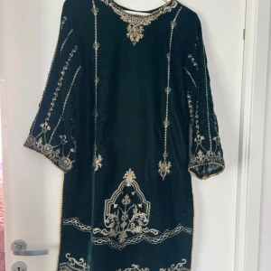 Mörkgrön klänning med broderier och tillbehör  - Unik mörkgrön klänning i sammet med vackra guldiga broderier och detaljer längs ärmar, halsringning och nederkant. Klänningen har trekvartsärmar och en rak passform. Perfekt för speciella tillfällen när du vill sticka ut. Har en i samma storlek fast mörkblå så tvek inte och ge ett meddelande. Kommer med sjalen och byxorna. 