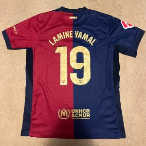 FC Barcelona Lamine Yamal matchtröja 2025/2025 - Säljer en officiell FC Barcelona fotbollströja med Lamine Yamal och nummer 19 på ryggen. Tröjan är röd och blå, har korta ärmar, guldiga detaljer och klubbmärke framtill. Materialet är lätt och andas, perfekt för match eller träning.