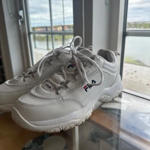 Vita sneakers från Fila - Säljer ett par klassiska vita sneakers från Fila med chunky sula och snörning. Skorna har Fila-logga på sidan, plösen och hälen. Perfekta för en sportig och trendig look.