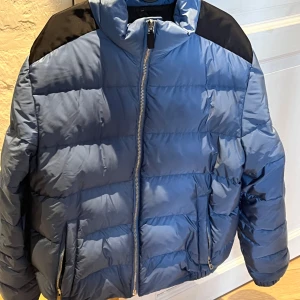 Blå pufferjacka från Prada - Säljer en blå pufferjacka från Prada med svarta axelpartier och hög krage. Jackan har dragkedja framtill och två fickor med dragkedja. Klassisk quiltad design som ger en modern look. Har Oxo två fickor inne och en på armen skick 9/10 använd fåtal gånger storlek M passar L med