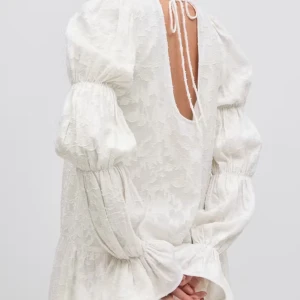 Vit blommig kortklänning med volangärm - Superfin vit kortklänning med subtilt blommönster och volangdetaljer på ärmarna. Klänningen har lång ärm, öppen rygg med knyt och en lös passform. Perfekt för sommarens tillfällen!