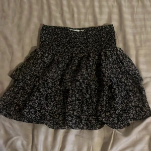 Blommig volangkjol från KappAhl - Supersöt kort volangkjol från KappAhl med svart botten och små blommor i vitt och ljusrosa. Kjolen har smock i midjan för extra bekväm passform och är perfekt för dig som gillar en feminin och lekfull stil. Själv rekommenderar jag att gå ner i en storlek eller två beroende på längg och hur du vill att den ska sitta. Själv är jag 165 cm lång och sitter som en mini kjol på mig.