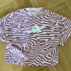 Rosa mönstrad t-shirt från Adidas - Säljer en rosa och ljusrosa t-shirt från Adidas med ett coolt zebramönster och vit logga framtill. T-shirten har korta ärmar och en lös passform, samt en snygg slits baktill för extra stilpoäng.