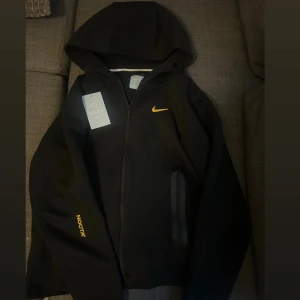 Nike NOCTA svart hoodiejacka XS/S - Svart hoodiejacka från Nike NOCTA med gul Swoosh på bröstet och NOCTA-tryck på ärmen. Jackan har hel dragkedja, huva och diskreta detaljer i gult på ryggen. Tillverkad i mjukt material, perfekt för en clean och sportig look. Står storlek M i, men passar mer som S/XS.