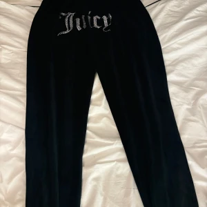 Svarta Juicy Couture mjukisbyxor med strass - Svarta mjukisbyxor från Juicy Couture i velour med glittrande strassdetaljer och stor 'Juicy'-text framtill. Byxorna har resår och snörning i midjan samt en liten strasslogga på ena fickan. Perfekta för en chill och trendig look. Storlek medium, knappt använda