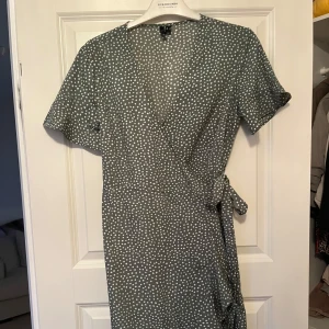 Prickig omlottklänning från Vero Moda - Grön omlottklänning från Vero Moda med vita prickar, kort ärm och knyt i midjan. Klänningen har volangdetalj nertill och v-ringad hals. Perfekt för dig som gillar en feminin och trendig look.