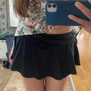 Svart kortkjol med vågig kant - Supersöt svart kortkjol med vågig nederkant och snygg drapering i midjan. Kjolen har en enkel och stilren look som passar perfekt till crop tops eller oversized skjortor. Materialet känns mjukt och följsamt mot huden.