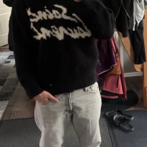 Svart stickad Saint Laurent tröja - Svart stickad tröja från Saint Laurent med vit, grafisk logga framtill. perfekt för dig som gillar streetstyle och vill sticka ut med en ikonisk look.