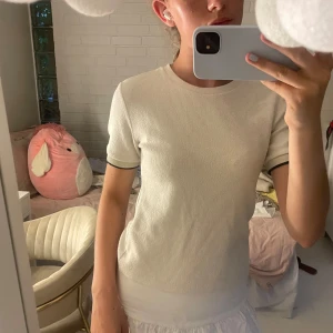 Vit ribbad topp med svarta detaljer - Säljer en stilren vit ribbad topp med korta ärmar och svarta tunna ränder längst ärmsluten. Toppen har en rund halsringning och är i ett mjukt, stretchigt material som sitter snyggt på kroppen. Perfekt för dig som gillar en clean och enkel look.