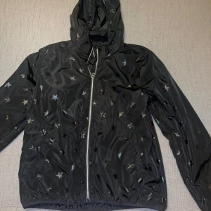 Svart jacka - Svart vindjacka från H&M med reflekterande stjärnmönster över hela jackan. Jackan har huva, dragkedja framtill och två fickor. Perfekt för dig som vill sticka ut lite extra när du är ute. Materialet är lätt och känns smidigt.