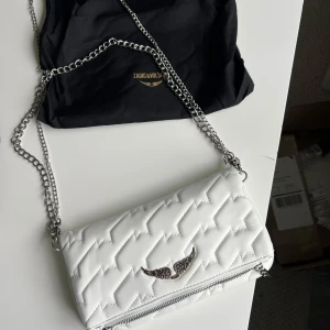 Vit quiltad axelväska Zadig & Voltaire - Supersnygg vit quiltad axelväska från Zadig & Voltaire med silverfärgad kedja och ikonisk vingdetalj framtill. Väskan har dragkedja och magnetstängning, samt ett rymligt innerfack. Perfekt för dig som vill ha en stilren och trendig accessoar. Aldrig använd 