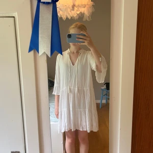 Vit kortklänning från Amisu - Supersöt vit kortklänning från Amisu med v-ringning och knytband framtill. Klänningen har trekvartsärmar och ett luftigt, flowy tyg som känns perfekt för varma dagar. Fina volangdetaljer och lätt struktur i materialet ger en chill och fräsch vibe.