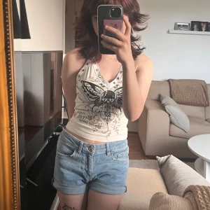 Vit halterneck linne med dödskallefjäril - Säljer ett vitt halterneck linne med coolt tryck av en dödskallefjäril och silvriga nitar längs v-ringningen. Toppen är croppad och har smala band som knyts i nacken. Perfekt för dig som gillar edgy och unik stil. Osäker på slt men passar xs, s, och möjligen m!