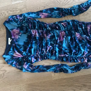 Blå och lila mönstrad klänning H&M - Säljer en cool kortklänning från H&M med blått och lila mönster. Klänningen har långa ärmar och är rynkad över hela kroppen för en snygg effekt. Perfekt för dig som gillar färg och vill sticka ut lite extra.