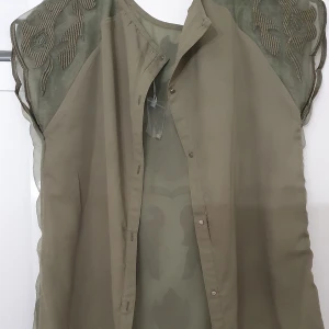 Blus - Olivgrön blus från Oxygene i storlek L. Transparent tyg med broderade detaljer och blommönster på axlar och rygg. Ärmlös modell med snygga knappar framtill och vågig nederkant. Perfekt för dig som gillar unika detaljer och en luftig känsla.