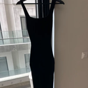 Svart långklänning med breda axelband, midi - En stilren svart långklänning med breda axelband och rak, tight passform. Klänningen har en klassisk, minimalistisk look och är ärmlös med fyrkantig ringning. Perfekt för dig som gillar enkel men elegant stil.