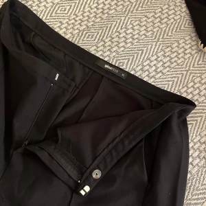 Svarta kostymbyxor från Gina Tricot XL - Snygga svarta kostymbyxor från Gina Tricot i storlek XL. Byxorna har klassisk rak passform med dragkedja och knapp framtill. Perfekta till en stilren look och enkla att matcha med olika toppar.