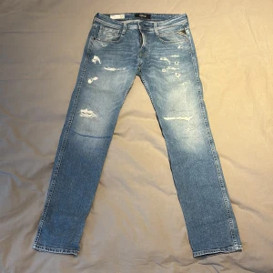 Replay Anbass ljusblå slim jeans - Snygga Replay Anbass jeans i ljusblå tvätt med slitningar och distressed detaljer. Slim fit modell med klassisk femficksdesign och dragkedjegylf. Tillverkade i ekologisk bomull för en skön känsla och modern look.