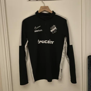 Svart AIK långärmad Nike träningströja - AIK fotbollströja från Nike i svart med vita detaljer längs sidor och ärmar. Långärmad modell med halv dragkedja och klubbmärke på bröstet. Sponsorer som Truecaller, Fabege och SVEA tryckta på fram- och baksidan. Perfekt för träning eller match.