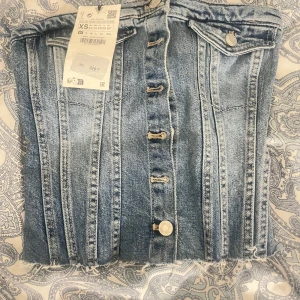 Jeans bandeau tubtopp med knappar XS - Säljer en snygg  tubtopp i jeans med silverfärgade knappar framtill och fransig nederkant. Toppen har dekorativa fickor upptill och markerade sömmar som ger en figurnära look. 