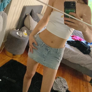 Ljusblå jeanskjol i kort modell - Säljer en ljusblå kort jeanskjol med klassisk femficksdesign och knapp framtill. Perfekt för dig som gillar en enkel och stilren look. Kjolen har en normal passform och är lätt att matcha med olika toppar.