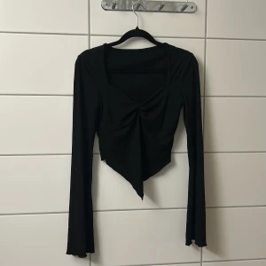 Svart långärmad croppad topp - Svart långärmad topp med v-ringning och twistad detalj framtill. Toppen är croppad med spetsig nederkant och har en tight passform. Perfekt för dig som gillar trendiga och stilrena plagg.