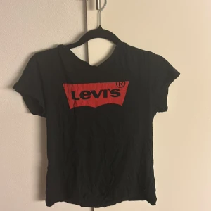Svart Levi's t-shirt med röd logga - Svart t-shirt från Levi's med klassisk röd logga framtill. T-shirten har korta ärmar och rund halsringning. Perfekt basic-plagg med streetkänsla och tydlig branding. Materialet känns mjukt och passar till många olika stilar.