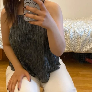 Glittrig svart linnetopp från Gina Tricot - Säljer en snygg glittrig svart linnetopp från Gina Tricot. Toppen har tunna axelband och ett rakt snitt med metallicskimrande tyg som verkligen sticker ut. Perfekt för dig som vill ha något extra till din outfit!