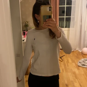 Vit långärmad tröja från Ralph Lauren - Vit långärmad tröja från Ralph Lauren med klassisk rund hals och den ikoniska blå broderade loggan på bröstet. Tröjan är i mjuk bomull och har en clean, enkel look som passar till allt. Perfekt för lager-på-lager under hösten eller våren.