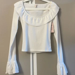 Vit blus med volang från Nelly - Supersöt vit blus från Nelly i storlek XXS. Blusen har en bred volang runt den vida ringningen och långa ärmar med volang vid ärmsluten. Materialet är lätt strukturerat och ger en fräsch känsla. Perfekt för dig som gillar romantiska detaljer och stilren look.