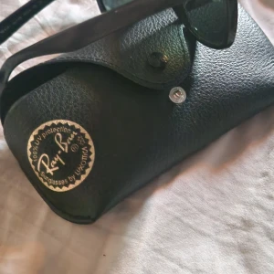 Svarta Ray-Ban solglasögon - Klassiska svarta Ray-Ban solglasögon med mörka glas och ikonisk form. Säljs med original svart fodral i skinnimitation med Ray-Ban-logga i guld. Perfekta för en cool och tidlös stil.