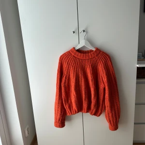 Orange stickad tröja från Envii - Supermysig orange stickad tröja från Envii med chunky ribbstickat mönster. Tröjan har en hög, rund halsringning och långa ärmar med breda muddar. Perfekt för lager-på-lager och ger en cool, avslappnad vibe till din outfit.