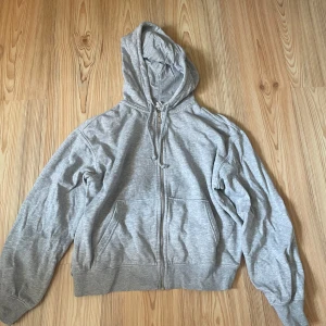Grå hoodie med dragkedja  - En basic grå hoodie från H&M med dragkedja framtill, huva med snörning och två stora fickor. Tillverkad i mjuk bomullsblandning, perfekt för chill dagar eller när du vill ha en enkel och clean look.