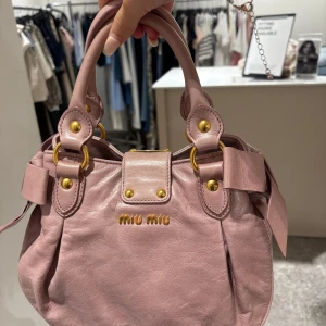 Rosa handväska från Miu Miu i skinn - Supersnygg rosa handväska från Miu Miu i mjukt skinn med guldiga metalldetaljer och dubbla handtag. Väskan har dekorativa sömmar och coola ringar i metall samt en diskret logga framtill. Perfekt för dig som vill ha en trendig och lyxig accessoar. Säljer för 4560kr❤️