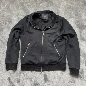 Svart zipjacka Limitato x Riocam - Svart zipjacka med coolt konstmotiv på ryggen, designad av Limitato i samarbete med Riocam. Jackan har två snedställda fickor med dragkedja, ribbade muddar och broderad logga på bröstet. Perfekt för dig som vill sticka ut med stil.