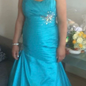 Turkos långklänning med paljettdetaljer - Säljer en turkos långklänning med glittrande paljettdetaljer vid midjan och höften. Klänningen har draperad överdel och figurnära passform upptill, med en vacker utsvängd kjol. Perfekt för bal eller festliga tillfällen.