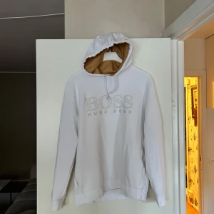 Vit hoodie från Hugo Boss - Snygg vit hoodie från Hugo Boss med stor BOSS-logga broderad på bröstet. Tröjan har huva med beige insida och snörning, samt långa ärmar och ribbade muddar. Perfekt för en clean och stilren look. Köptes på NK för 2399kr