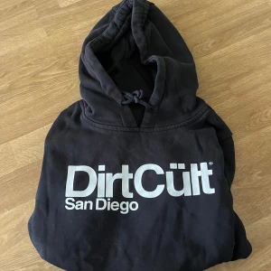 Svart Dirt Cult hoodie med tryck - Svart hoodie från Dirt Cult med stor vit logga och texten San Diego på bröstet. Klassisk huva med snörning och mjukt material som känns skönt mot huden. Perfekt för dig som gillar streetwear och vill sticka ut med en cool print.