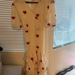 Långklänning med röda hjärtan - Romantisk beige långklänning  med röda hjärtan, puffärmar och omlottliv. Tjejen i bilden är ca 167cm lång. Klänningen har volangdetaljer nertill och knytband i midjan. 