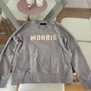 Grå sweatshirt från Morris - Säljer en grå sweatshirt från Morris med broderad logga i vitt framtill. Tröjan har rund halsringning, långa ärmar och ribbade muddar vid ärmslut och nederkant. Perfekt för en chill och stilren look. Säljer pga för liten. Kommer att tvättas innan leverans.