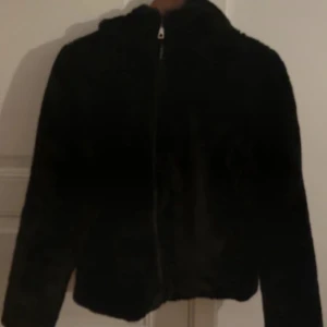 Svart pälsjacka från Vero Moda - Mysig svart pälsjacka från Vero Moda med huva och dragkedja framtill. Jackan har en fluffig känsla och är perfekt för kalla dagar. Enkel och stilren design som passar till många outfits.