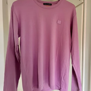 Ljuslila långärmad tröja Acne Studios - Säljer en stilren ljuslila långärmad tröja från Acne Studios i 100% bomull. Tröjan har rund halsringning och den ikoniska Face-loggan broderad på bröstet. Perfekt för dig som gillar minimalistisk och trendig design.