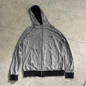 Grå hoodie från Hugo Boss med dragkedja - Snygg grå hoodie från Hugo Boss med svart foder i huvan och svarta muddar. Tröjan har hel dragkedja framtill, två fickor och diskret Boss-logga på bröstet.