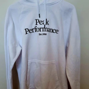 Vit hoodie från Peak Performance - Säljer en vit hoodie från Peak Performance med svart broderad logga på bröstet. Tröjan har huva med snörning, stor magficka och långa ärmar. Perfekt för dig som gillar en clean och sportig stil.
